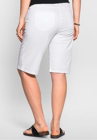Persona vistiendo pantalones cortos blancos hasta la rodilla, camiseta negra, sandalias negras tipo slider y una pulsera, de pie contra un fondo liso.