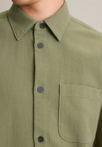 Olive green button-up shirt with a textured fabric, pointed collar, black buttons, and a chest pocket on the left side.