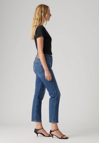 Camiseta negra ajustada, vaqueros de tiro alto azules con un dobladillo recortado y sandalias negras con tiras. Logo notable de Levi's en el bolsillo trasero.