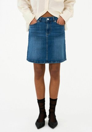 Mujer que lleva una falda vaquera azul con bolsillos delanteros, calcetines negros transparentes, tacones negros de punta y camisa crema con botones.