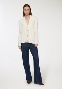 Cardigan senza maniche lavorato a maglia color avorio con scollatura a V profonda, adornato con grandi bottoni rotondi, abbinato a jeans a zampa d'elefante blu e scarpe marroni a punta.