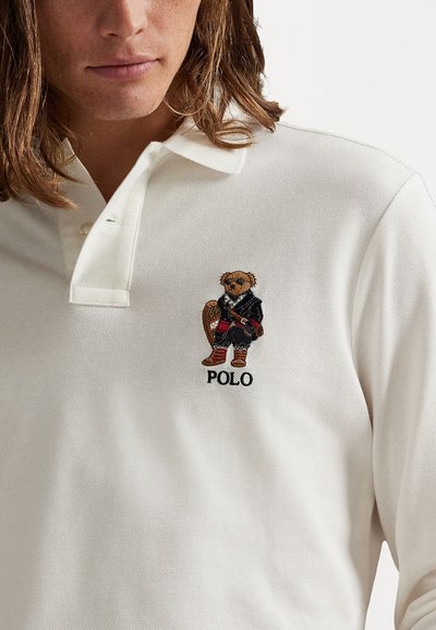 Bílá polo košile s vyšívaným medvědem v černé bundě a červených kalhotách, s textem "POLO" pod designem.