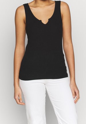 Kobieta ubrana w bezrękawnik w kolorze czarnym z dekoltem wciętym i białe spodnie, stojąca na jednolitym jasnym tle.