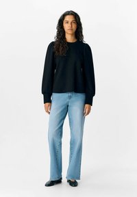 Pull en noir à manches bouffantes longues, assorti à un jean droit bleu clair et des ballerines noires, sur fond blanc uni.