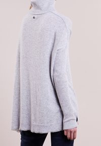 Grå stickad polotröja med ribbad textur, sidslitsar och en liten logotyp på baksidan. Oversized passform med långa ärmar.