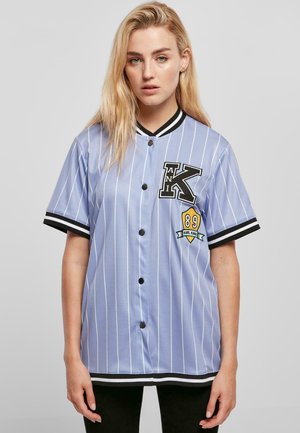 Lyseblå baseballtrøje med lodrette hvide striber, sorte accenter på ærmerne og kraven, der har et stort sort "K" og et emblem med "89."