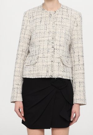 Woman wearing a cream and black tweed jacket with pearl buttons over a black skirt with a floral fabric detail on the waist.