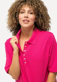 Roze polo met korte mouwen en een knoopsluiting. Zachte textuur en kraagdetails zichtbaar. Model houdt de kraag omhoog.