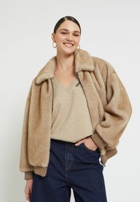 Beige faux fur bomber jacket with a large collar, ribbed cuffs, and a zip closure. Paired with a light beige V-neck top and dark denim jeans.