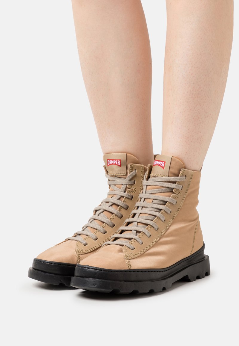 Beige hohe Schnürstiefel mit dicken schwarzen Sohlen, die auf bloßen Beinen vor einem weißen Hintergrund getragen werden.