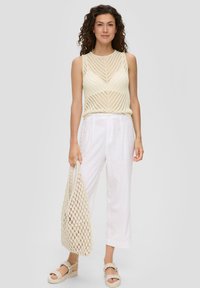 Crème gebreide tanktop met een chevronpatroon, gecombineerd met witte linnen broek. Het model houdt een beige gehaakte tas vast en draagt geweven sandalen.