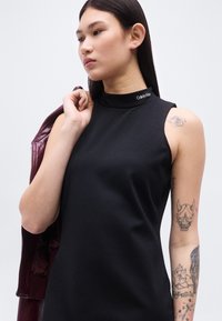 Νεαρή γυναίκα με μακριά σκούρα μαλλιά φοράει μαύρο αμάνικο τοπ Calvin Klein, κρατώντας μπορντό σακάκι στον ώμο, με πολλά τατουάζ στα μπράτσα.