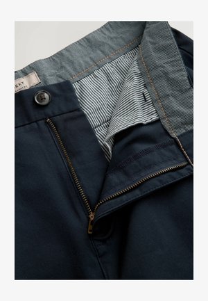 Tummansinisiä suoria housuja, joissa on vetoketjukiinnitys, nappisulku ja raidoitettu kangaspäällyste vyötäröllä. Esillä ompelutikkaus.