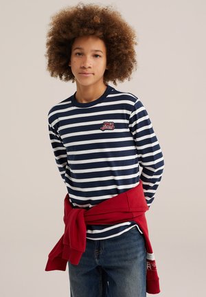 Jeune personne aux cheveux bouclés portant un tee-shirt à manches longues rayé bleu marine et blanc et un pull rouge noué autour de la taille.