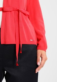 Cárdigan de coral con mangas largas, cintura con cinturón y puños acanalados. Llevado sobre pantalones negros texturizados, con un acento de logo metálico.