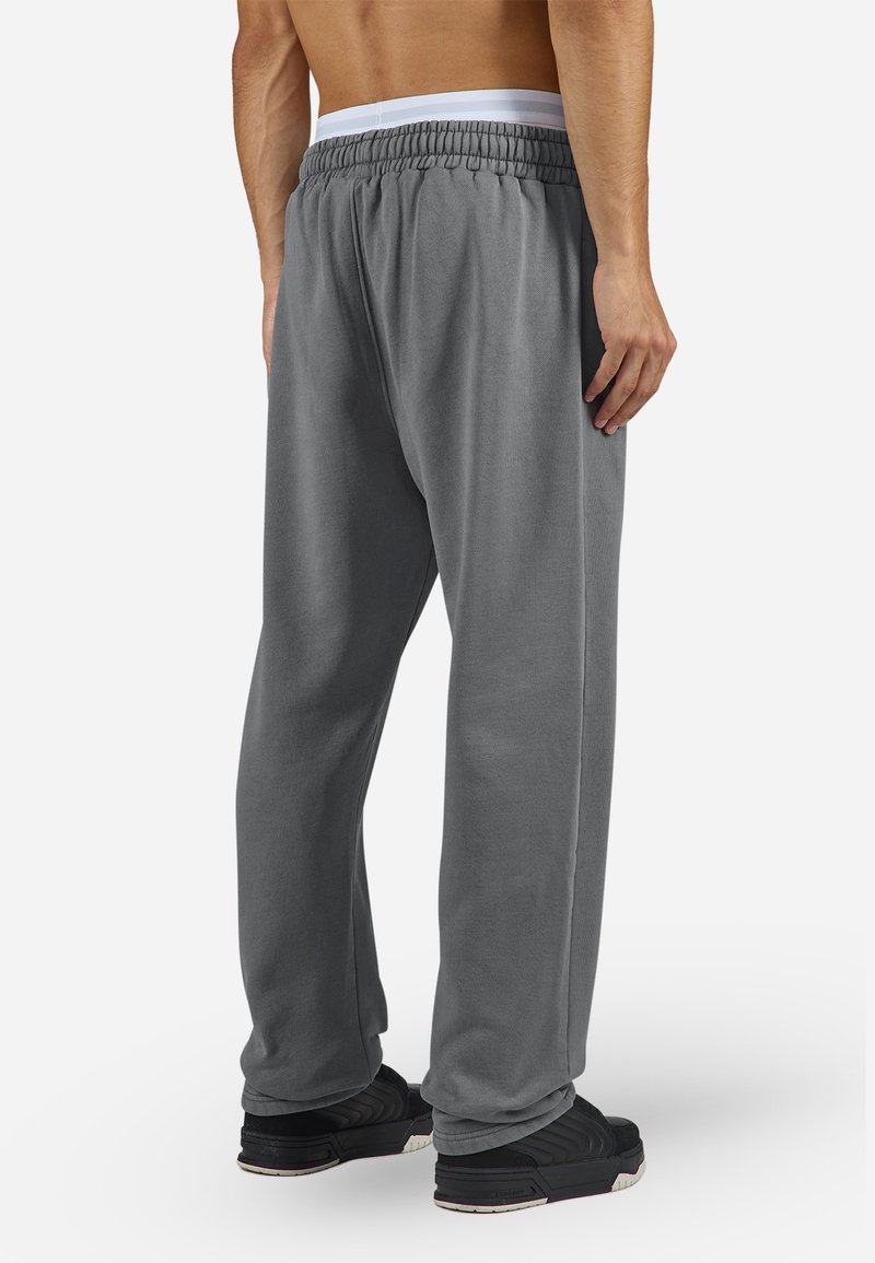 Graue Jogginghosen mit einem elastischen Bund und weißem Innenfutter, mit entspannter Passform und glatter Textur, getragen mit schwarzen Sportschuhen.