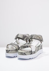 Sandalové klíny se šedým hadím vzorem, nastavitelné popruhy, bílé podrážky a kovovým logem Calvin Klein na popruhu kolem kotníku.