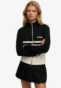 Czarna bluza rozpinana na zamek z kremowymi paskami akcentuj�ącymi i logo "SUPERDRY". Posiada wysoki kołnierz i ściągacze na rękawach. Noszona z plisowaną czarną spódnicą.