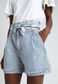 Niebiesko-białe szorty w paski z wysokim stanem, wiązane w talii, z plisowanym przodem i bocznymi kieszeniami. Wykonane z lekkiej tkaniny.