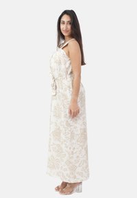 Frau, die vor einem einfachen weißen Hintergrund in einem cremefarbenen, floralem Maxikleid mit Taillengürtel steht. Sie hat langes, dunkles Haar und trägt klare, hochhackige Sandalen.