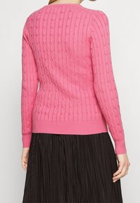 Maglione a trecce di color rosa brillante, con maniche lunghe, scollatura rotonda e orlo aderente; texture dettagliata visibile su tutto il capo.