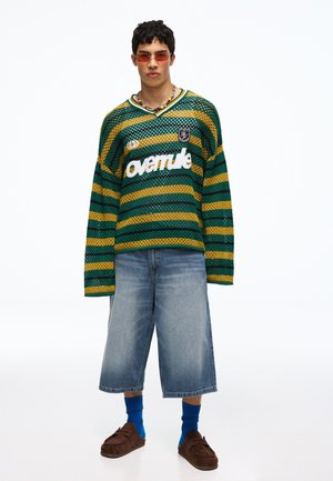 Jeune homme portant un pull en maille rayé vert et jaune, un long short en jean, des chaussettes bleues, des chaussures marron, un collier coloré en perles et des lunettes de soleil teintées orange.