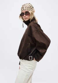 Γυναίκα που φοράει σκούρο καφέ φαρδιά ζακέτα, άσπρο παντελόνι, μαντήλι με λεοπάρ μοτίβο στο κεφάλι και μαύρα γυαλιά ηλίου, ποζάροντας μπροστά σε απλό ανοιχτό φόντο.