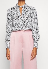 Blusa a fiori bianca e nera con dettaglio a nodo. Abbinata a pantaloni a vita alta e larghi di colore rosa chiaro. Tessuto dalla consistenza morbida.
