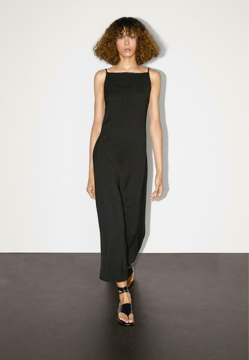 Svart ärmlös jumpsuit med fyrkantig halsringning, flödande design, tillverkad av ett texturerat tyg, matchad med svarta platta sandaler.