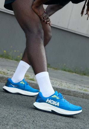 Zapatillas de running HOKA azules con parte superior de malla, media suela blanca y acentos amarillos. Estilizadas con calcetines blancos y situadas sobre un fondo gris.