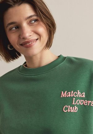 Zelená mikina z mäkkej tkaniny, s ružovým vyšitým textom "Matcha Lovers Club." Žebrovaný výstrih a pohodlný strih.
