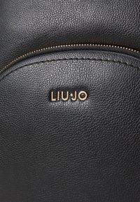 Musta teksturoidulla nahkabag, jossa on kultasävyinen vetoketju ja "LIU·JO" -logo. Sileä pinta, jolla on tyylik�äs, pyöreä muotoilu.