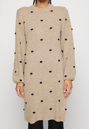 Beige gebreide sweaterjurk met ballonmouwen en verspreide zwarte pompon stippen, gedragen over een zwarte broek.