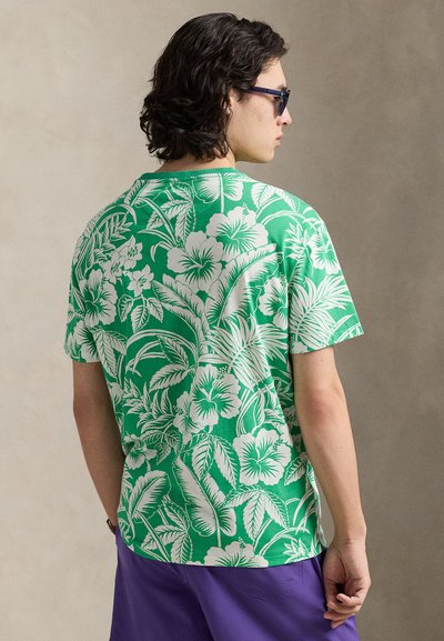 Πράσινο κοντομάνικο μπλουζάκι με λευκό σχέδιο φλοράλ και φύλλα, κατασκευασμένο από βαμβάκι. Φοράει μωβ σορτς, ορατό από πίσω.