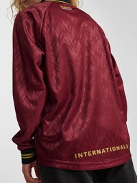 Isik, kes kannab tumesinist (maroon) mustrilist spordijopet kollaste tähtedega "INTERNATIONALS" allosas ning mustade varrukaservadega, millel on kollased triibud.