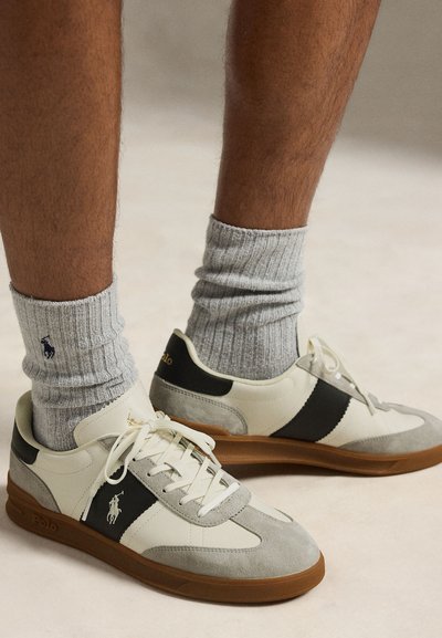 Picioare purtând șosete groase gri și adidași albi, negri și gri, cu tălpi maro, cu un logo de cal pe lateral.