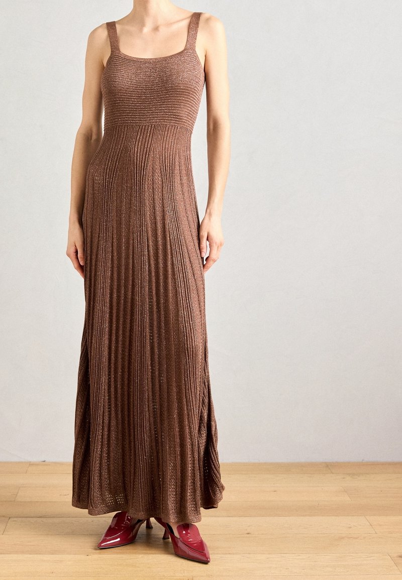 Robe longue en maille marron avec un corsage ajusté, un décolleté carré et une jupe plissée. Associée à des talons rouges à bouts pointus sur un sol en bois.