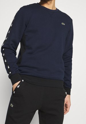 Man draagt een marineblauwe sweatshirt en zwarte joggingbroek met witte logodetails op mouwen en zakken, staat met handen in de zakken.