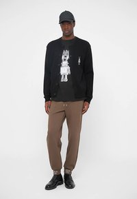 Svart stickad kofta över en grå t-shirt med robotmotiv, kombinerad med bruna sweatpants och svarta skor.