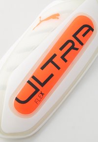 Hvit atletisk leggbeskytter med en oransje rektangulær aksent og svart tekst som sier "ULTRA FLEX." Har en Puma-logo i oransje.