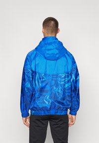 Giacca a vento blu con cappuccio caratterizzata da un motivo a gradiente. Il materiale è lucido e liscio, con polsini e orlo elastici.