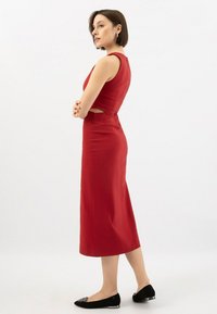 Donna in un vestito midi rosso senza maniche con ritagli laterali, in piedi di lato con le braccia incrociate, indossa delle ballerine nere con dettagli argentati.