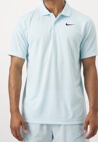 Jasnoniebieska koszulka polo z przewiewnej tkaniny, z czarnym logo Nike na lewej piersi, krótkimi rękawami i klasycznym kołnierzykiem.