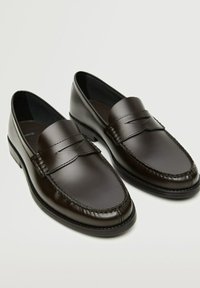 Pari ki hi-polish kiye hue kaale bhure chamde ke penny loafers jismein silayi ka kaam hai aur ek neecha heel hai, ek halka background par.