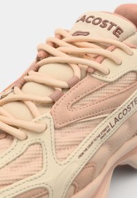 Tenisky Lacoste s vrškem z prodyšné tkaniny a semiše v jemné růžové a krémové barvě, se výrazným logem a texturovanými tkaničkami v odpovídající barevné kombinaci.