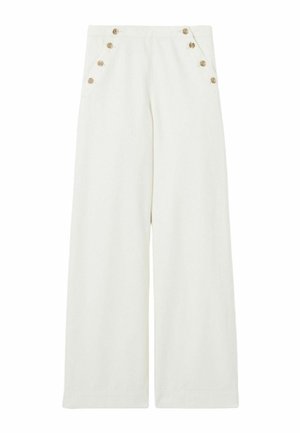 Jeune femme portant une chemise rayée bordeaux, un pantalon blanc large avec des boutons dorés, un chapeau bob rayé, tenant un sac tressé avec des accents en cuir.