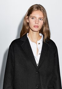 Czarny płaszcz z mieszanki wełny z klapami, z guzikami z przodu i luźnym krojem, założony na biały koszulę z akcentami złotych guzików.