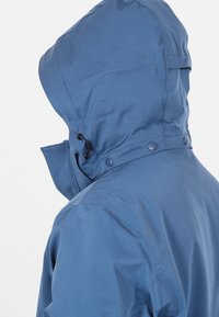 Person trägt eine blaue wasserdichte Jacke mit verstellbarer Kapuze und Knopfdetails vor einem einfarbigen weißen Hintergrund.