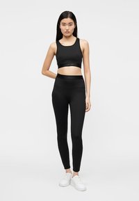 Brassière de sport noire et leggings en matière élastique. Les deux comportent une ceinture avec un logo discret. Assortis à des chaussures de sport blanches.