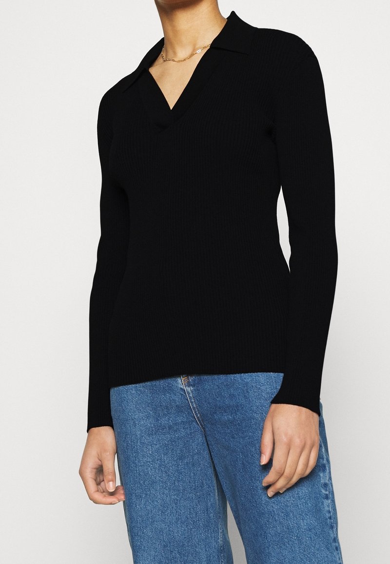 Femme portant un pull noir à manches longues avec col en V et un col, ainsi qu'un jean bleu, vue du cou jusqu'aux hanches sur un fond uni.